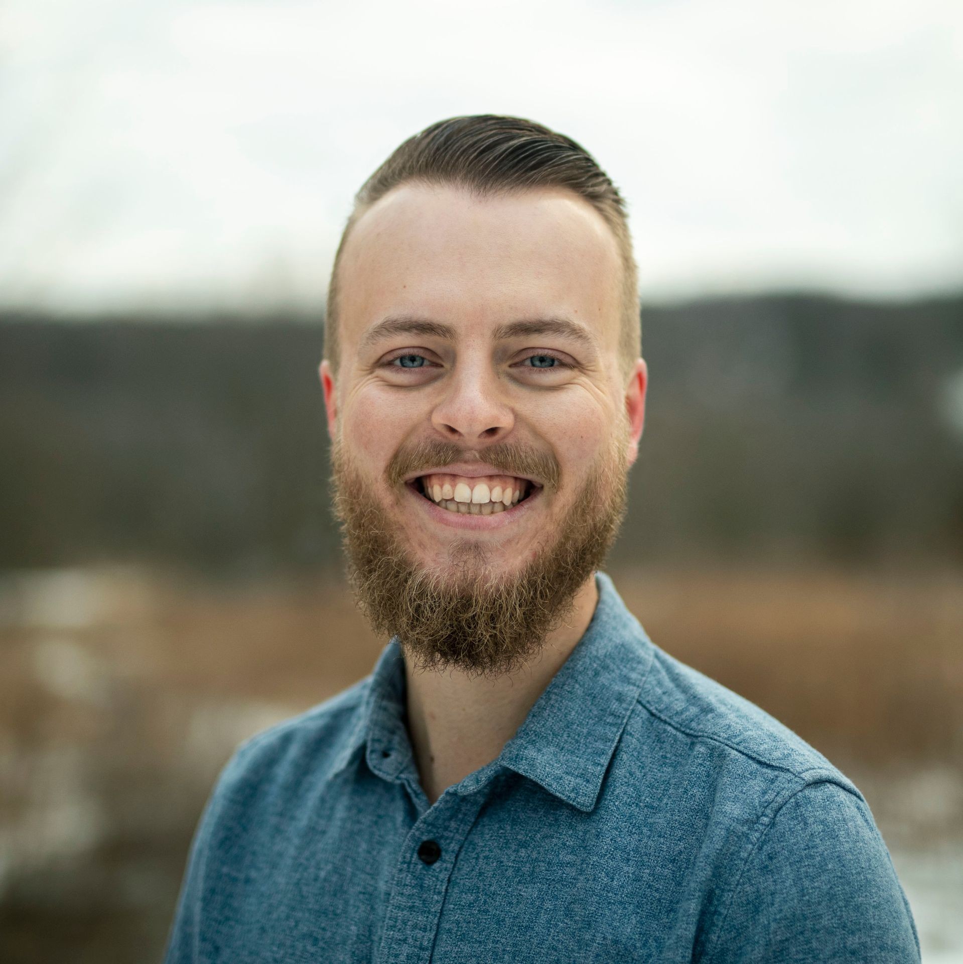 Person wearing a blue button-up shirt stands outdoors with a blurred natural background.